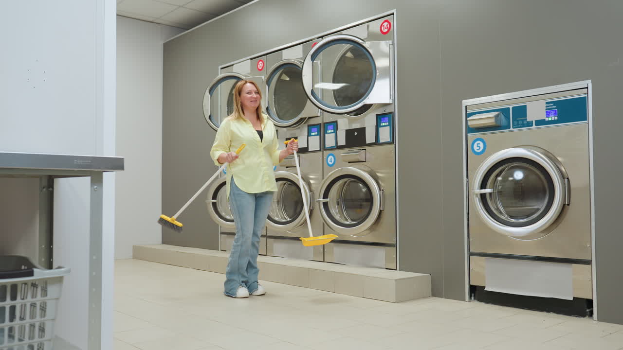 Happy laundromat worker holding broom and dustpan chats with unseen customer, smiling beside stainless industrial washers and dryers, showing cleaning routine, bright tiled interior, maintenance vibe