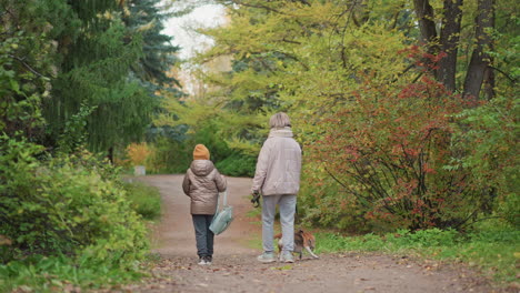 dog restrained by owner hand as it tries to dash into bush, kid adjusts bag strap while walking home on leafy forest path, warm jackets and scarf visible, vibrant autumn foliage framing scene