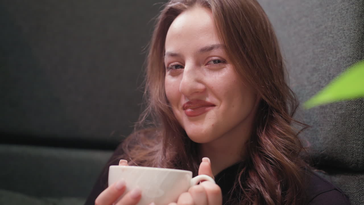 Lady holds white cup with both hands and gently sips warm latte with eyes closed, expressing peace and contentment. Soft lighting and cozy ambiance create soothing atmosphere
