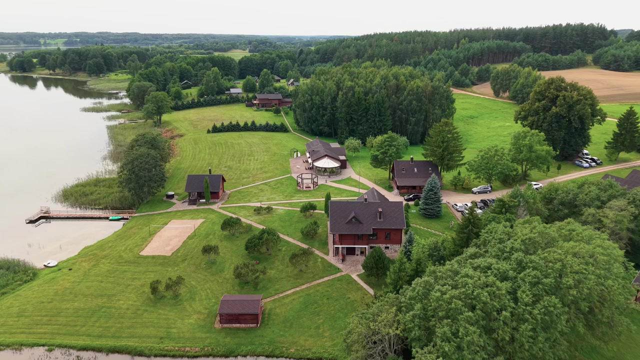 Drone-video tallentaa laajat näkymät rauhallisesta järvenrantaisesta maaseutukohteesta, jossa on viehättäviä puumökkejä, reheviä nurmikoita, puita, laituri ja tyyni ranta pehmeässä päivänvalossa.
