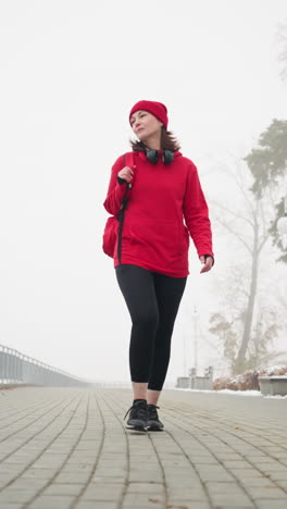 woman in red winter jacket and head warmer walks along snow-dusted paved pathway in foggy atmosphere with headphones resting around neck, surrounded by leafless trees and benches