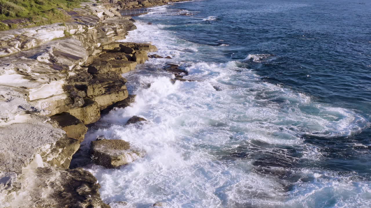 While fringed waves crashing onto ocean rocks POV drone moving slowly forward along cliff face ocean pool in background Coogee Beach Sydney Australia