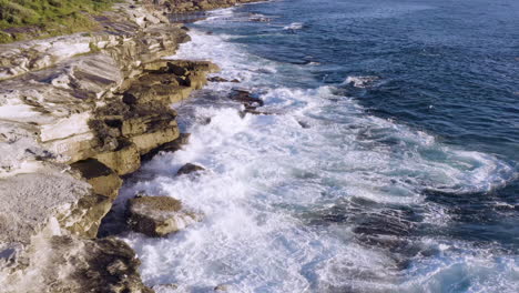 Mientras-Las-Olas-Con-Flecos-Chocan-Contra-Las-Rocas-Del-Océano,-El-Dron-Pov-Se-Mueve-Lentamente-Hacia-Adelante-A-Lo-Largo-De-La-Cara-Del-Acantilado,-La-Piscina-Del-Océano-En-El-Fondo,-La-Playa-De-Coogee,-Sydney,-Australia.