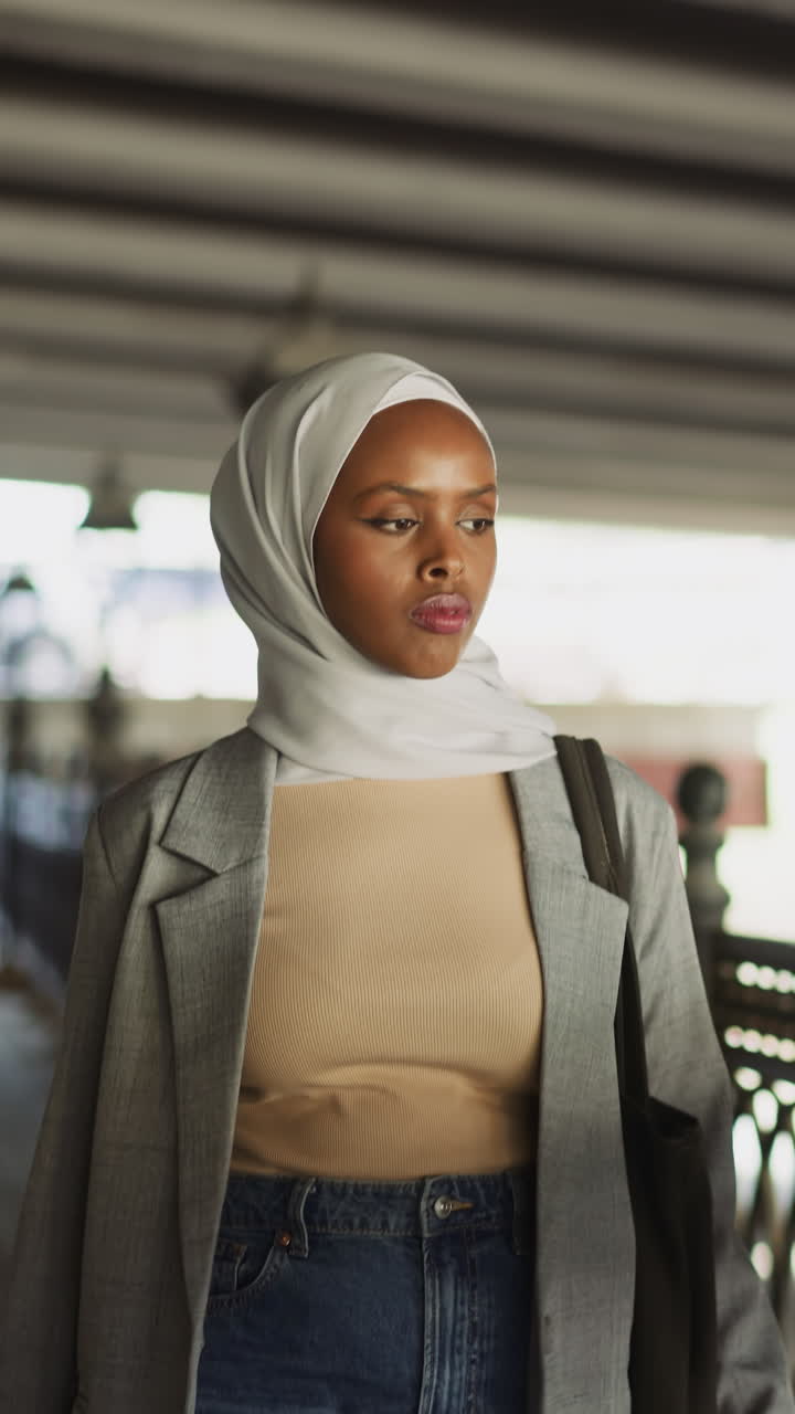 Stylish African-American woman with headscarf walks along waterfront railing under concrete bridge. Muslimah enjoys promenade in town. Arabian lady lifestyle