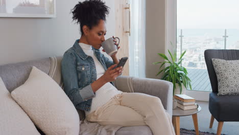 Mujer-Joven-Feliz-Usando-Un-Teléfono-Inteligente-Navegando-En-Línea-Leyendo-Mensajes-De-Redes-Sociales-Disfrutando-De-Un-Estilo-De-Vida-Cómodo-Bebiendo-Café-Relajándose-En-El-Sofá-En-Casa