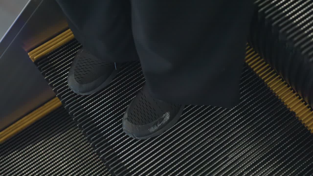 Leg view of person wearing black shoes and loose pants standing on moving escalator, showcasing modern urban transit and footwear in indoor public environment with visible motion lines