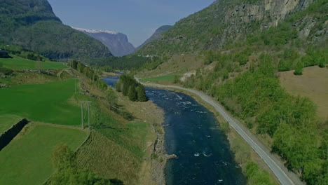 Aerial-backwards-shot-of-blue-river-surrounded-by-greened-mountains-and-snowy-peak-in-background---Summer-time-in-Norway,Europe