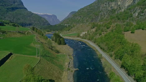 aerial backwards shot of blue river surrounded by greened mountains and snowy peak in background - summer time in norway,europe