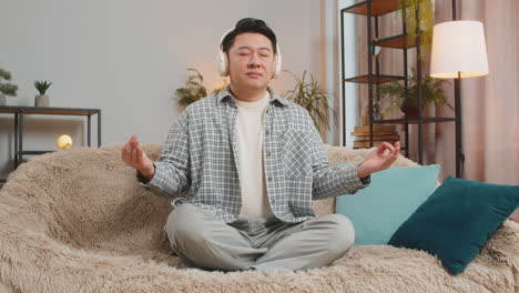 무선 헤드폰을 착용한 행복하고 편안한 젊은이가 집에서 소파에서 무드라 제스처로 깊은 숨을 쉬고 있습니다.