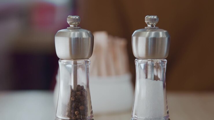 Close up of hand placing and adjusting salt shakers on table. pepper grinder contains whole peppercorns while adjacent container holds granulated salt. Soft lighting highlights clear glass
