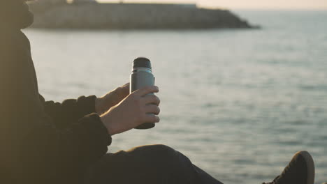 el hombre sentado en la playa abre el termo, vierte una bebida caliente en su taza, la bebe, la cierra, la llamarada de la hora dorada el sol golpea su espalda