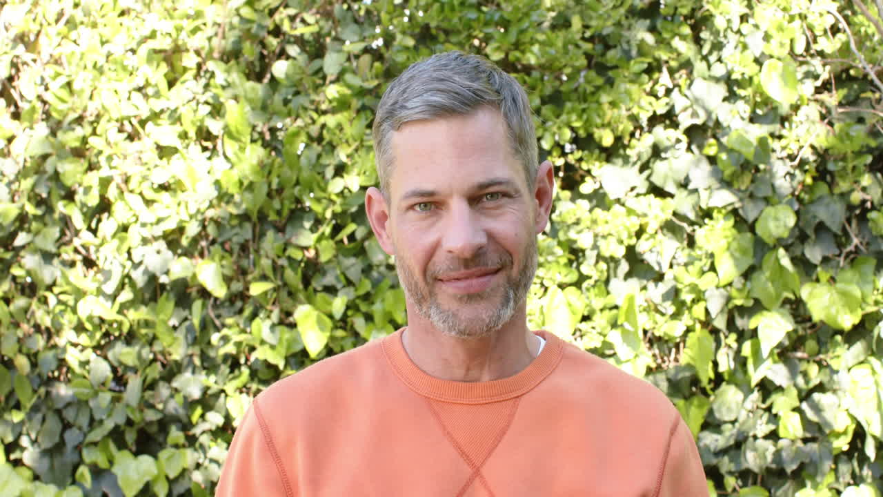 Homem caucasiano feliz com blusa laranja em pé e sorrindo em jardim ensolarado.