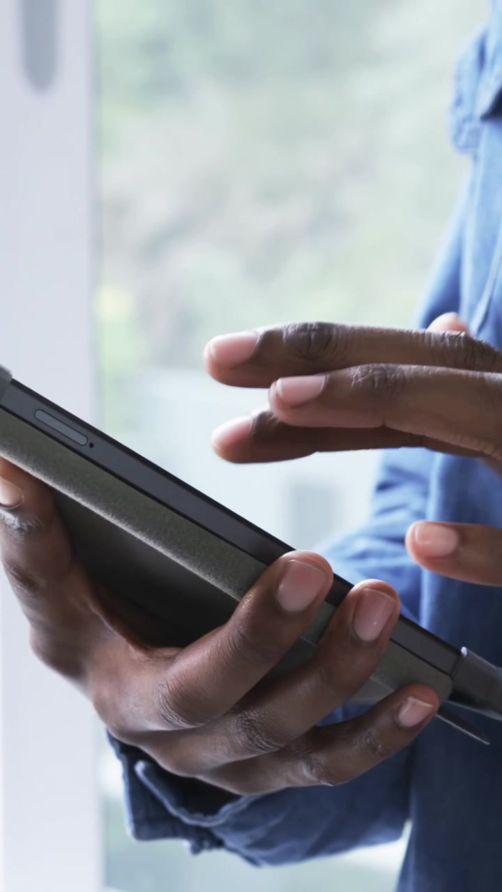 Vertical video: Tapping man wearing denim shirt swiping tablet near window, hands adjusting grip