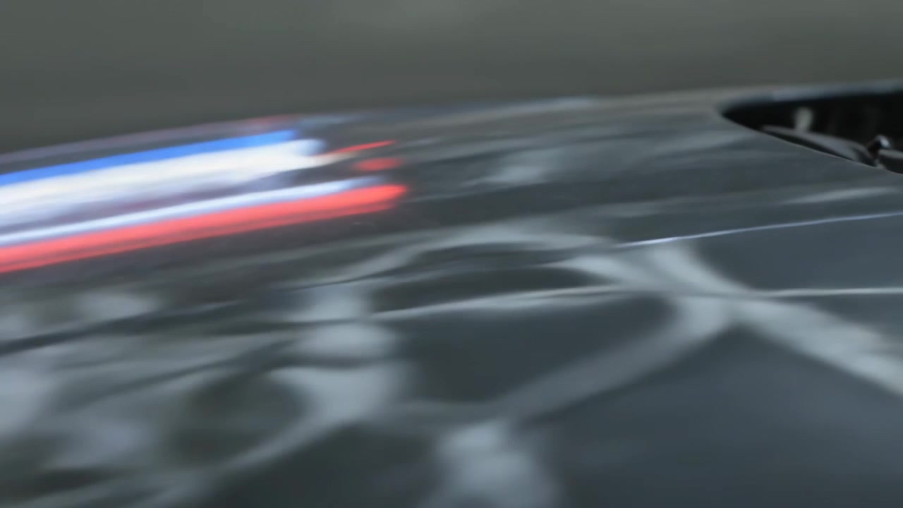 Fejende lys bevæger sig fra venstre mod højre, bilens motorhjelmsudluftning reflekterer rød-blå-hvide striber i et dæmpet studie.