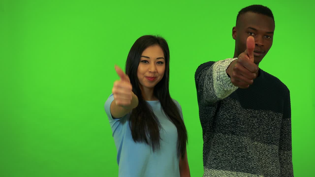 un joven negro y una joven asiática sonríen y muestran los pulgares hacia la cámara - estudio de pantalla verde