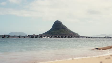 slow motion shot of Chinamans Hat Mokoli&#039;i in Oahu Hawaii with small waves crashing on the beach in the foreground