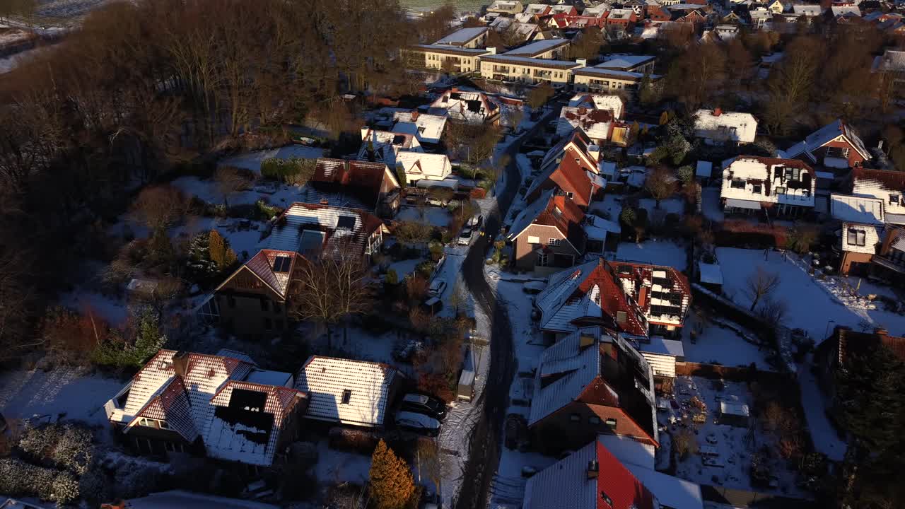 Letecký pohled na zasněženou německou obytnou čtvrť při západu slunce, kde teplé zlatavé světlo osvětluje střechy a úzké ulice. Klidná zimní atmosféra během malebného přeletu dronu.