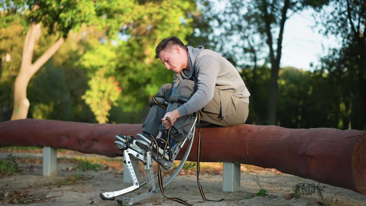짧은 갈색 머리카락을 가진 젊은 남자가 공원 같은 환경에서 큰 나무에 앉아 있으며, 밝은 회색 스웨트 셔츠와 카키 바지를 입고, 집중력으로 스 스트을 조정하는 데 집중하고 있습니다.