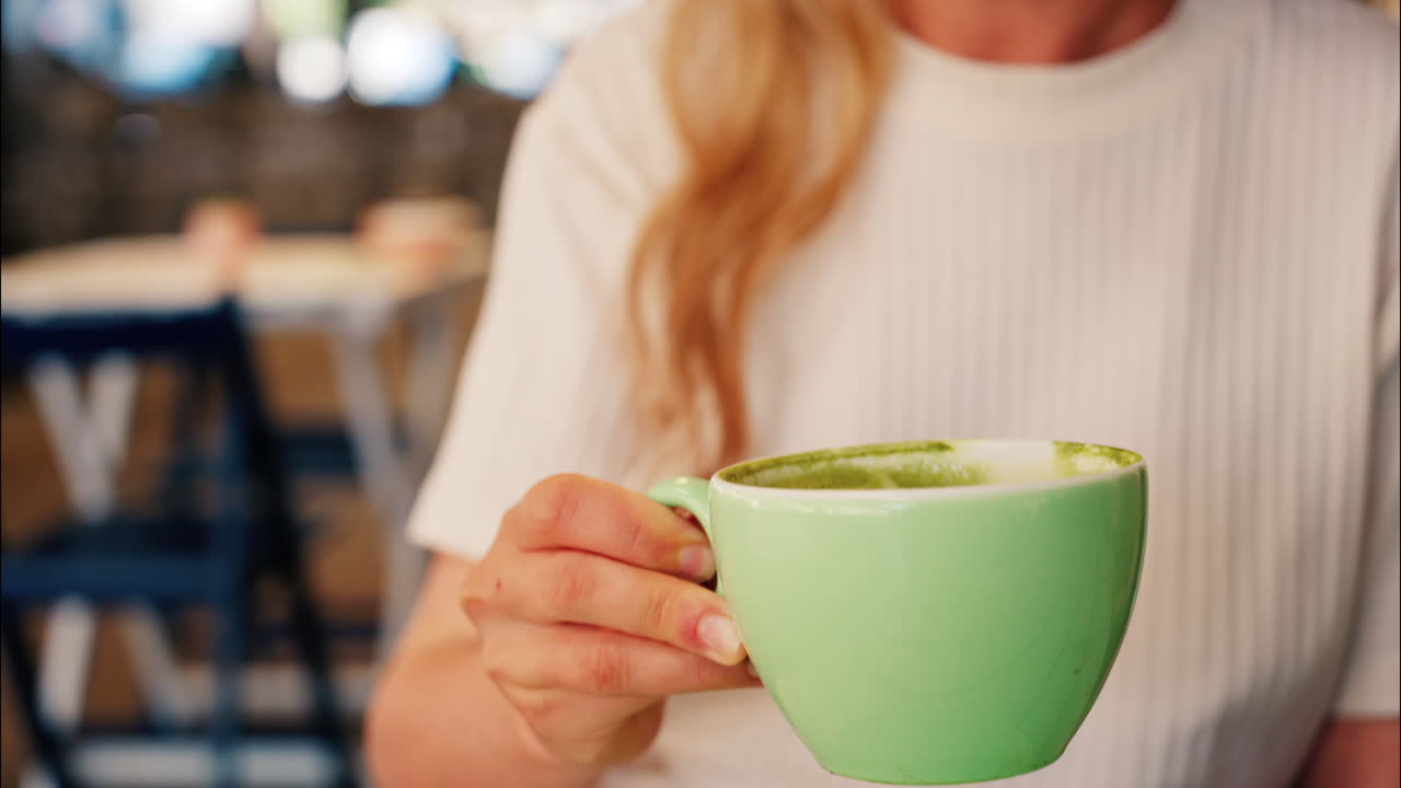 Close up of a blonde woman in a white T-shirt drinking a matcha latte out of a green cup at a terrace