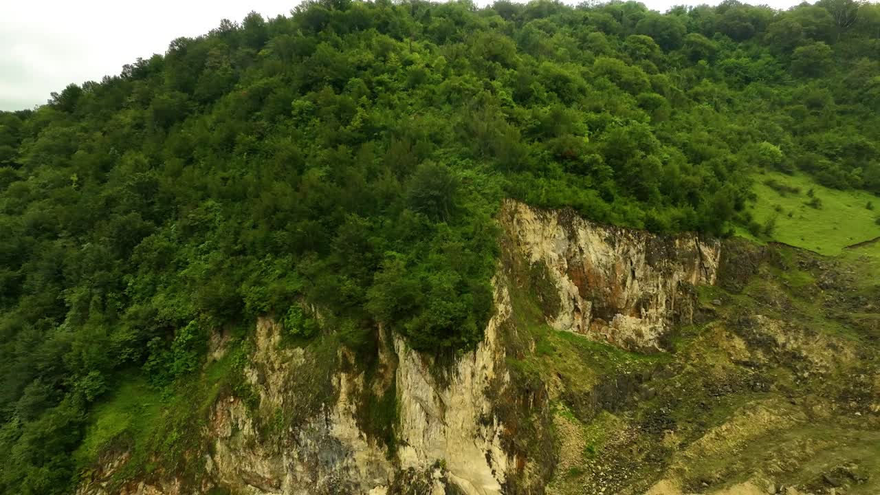 Aerial view of a steep rocky cliff covered in vibrant green vegetation, rising above a natural valley with a mix of stone and soil textures under soft daylight