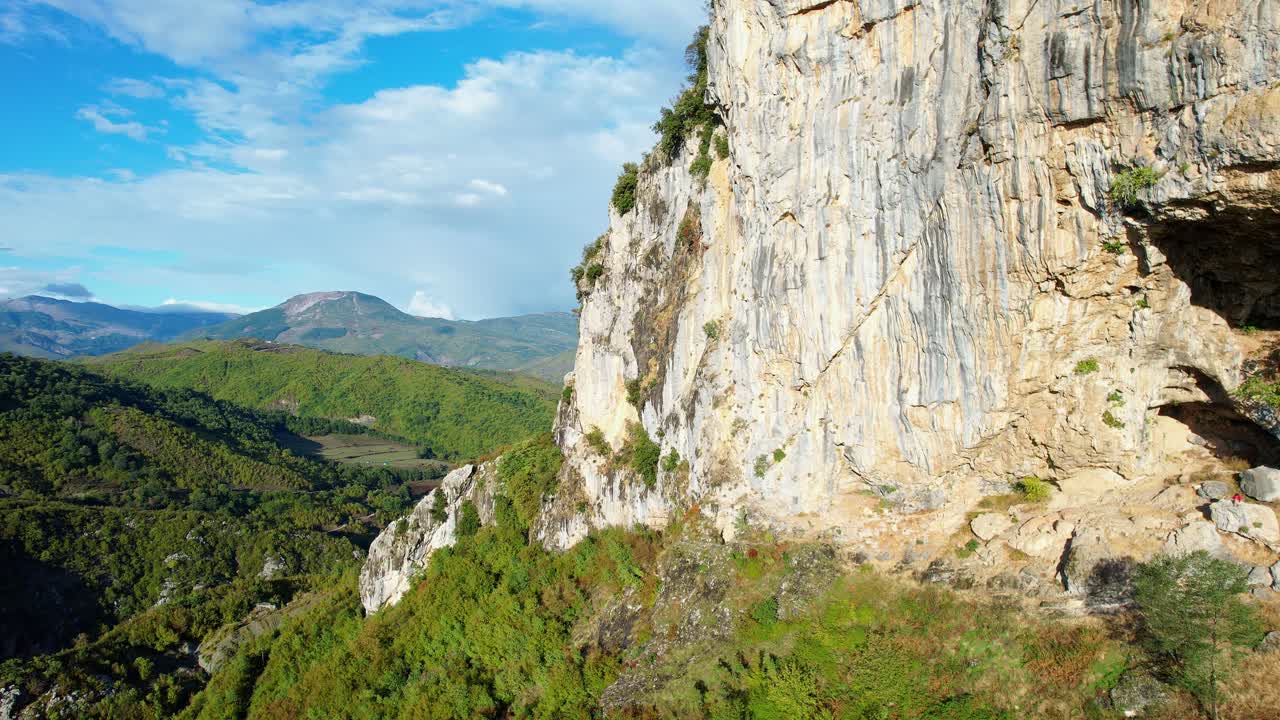 Side view (upward tilt-slow pan) of the Krrabë cliff in Albania under a blue sky. The shot highlights the rock texture and the stunning green, mountainous valley landscape
