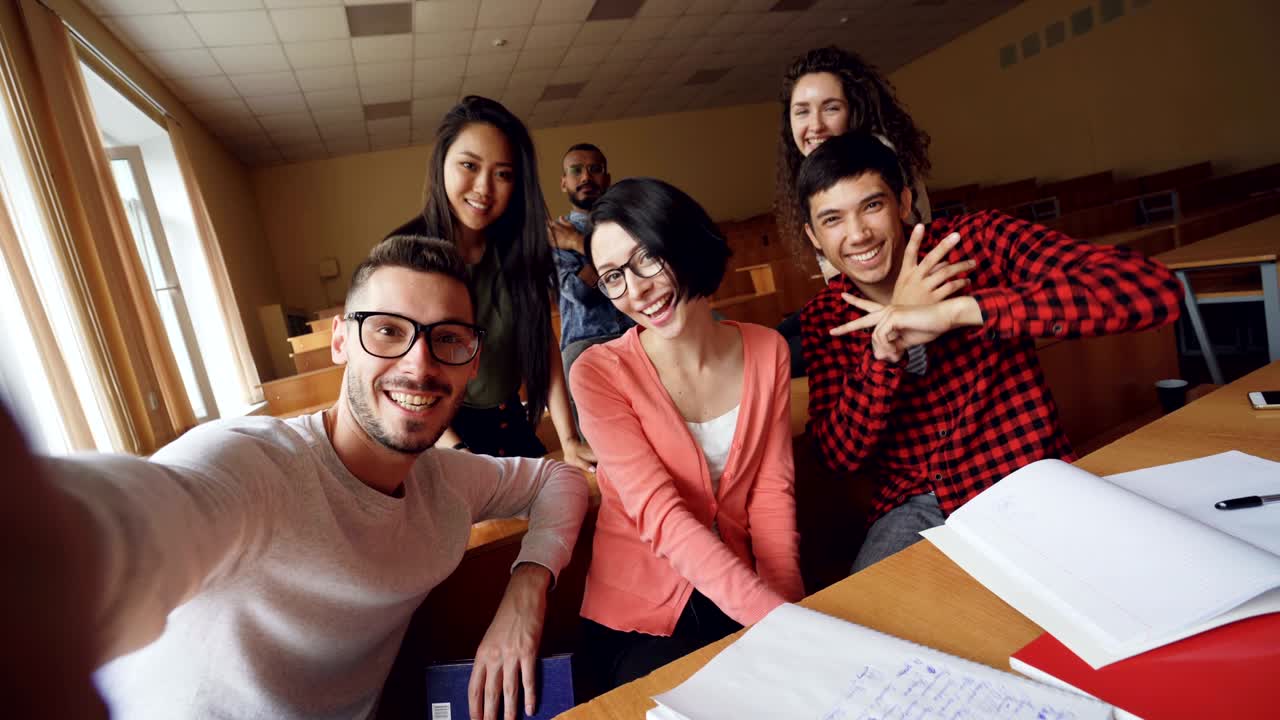 punto de vista de un grupo multiétnico de amigos tomando selfies en el aula mirando a la cámara, posando con gestos de la mano y sonriendo. tecnología moderna y concepto de milenios.