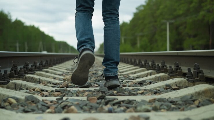 A close-up view of a man's legs, dressed in jeans and canvas shoes, walking alone on rocky railway tracks surrounded by dense trees and electric poles