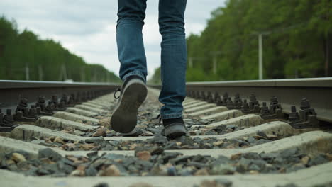 a close-up view of a man's legs, dressed in jeans and canvas shoes, walking alone on rocky railway tracks surrounded by dense trees and electric poles