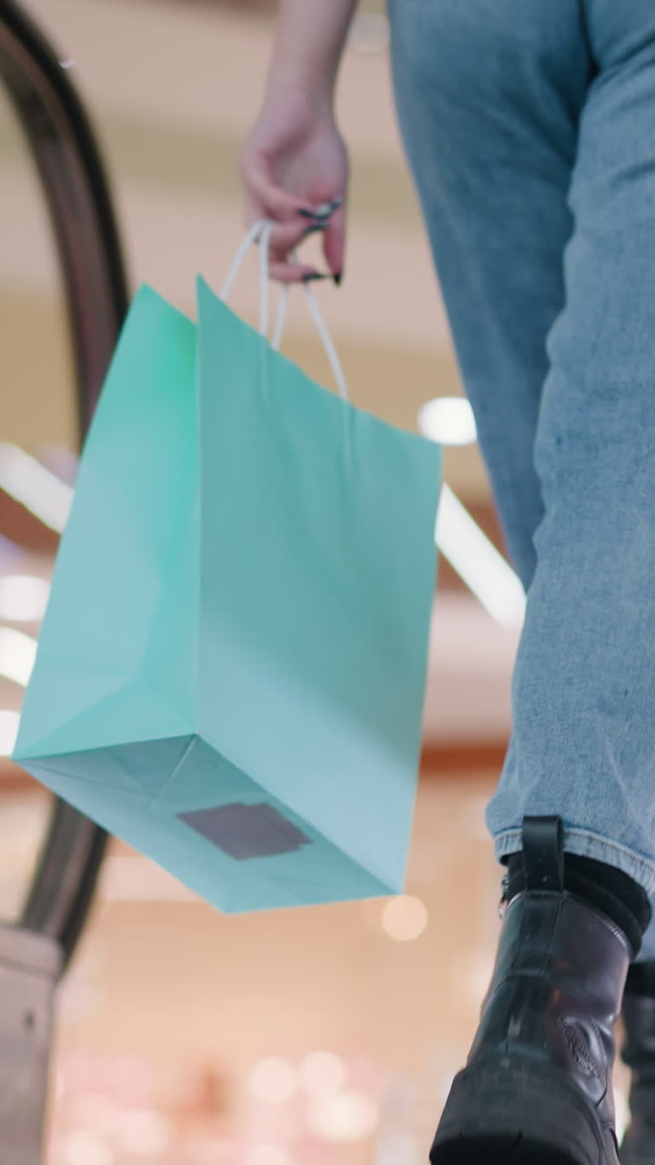 Leg view of woman with two shopping bags on moving escalator in mall, steps off onto brown rug with blurred background of mall's interior