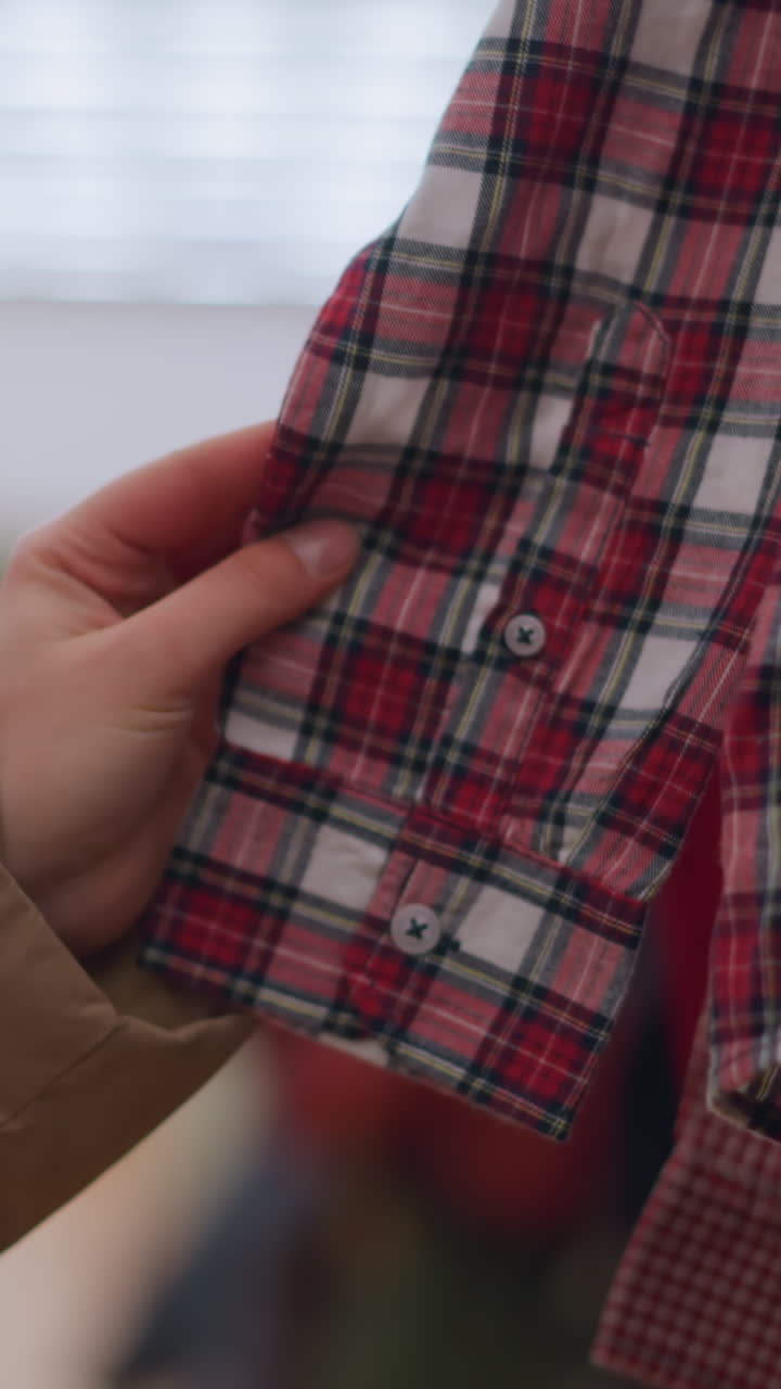 toma de primer plano de una mano con una chaqueta marrón claro, tocando suavemente camisas a cuadros colgando de un estante de ropa. la cara de la persona no es visible