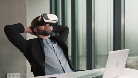 an relaxes by leaning back in his chair in the office, wearing virtual reality glasses and working on his laptop