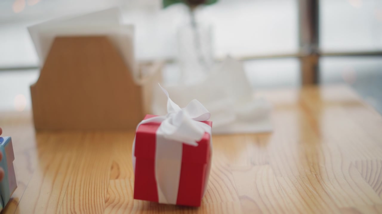 Child hands reach toward small wrapped gift on wooden tabletop while one hand holds crumpled tissue, with subtle motion, soft daylight, and cozy home setting suggesting recent handling or unwrapping