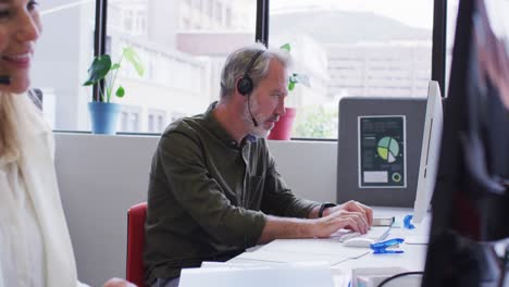 Caucasian-businessman-sitting-using-computer-talking-on-phone-headset-in-office