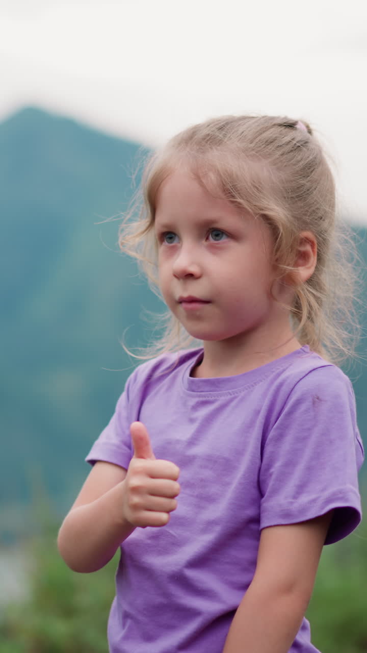 una niña linda con una trenza rubia muestra un gesto de pulgar hacia arriba admirando el valle verde con curvas río tranquilo en las montañas pie de primer plano en cámara lenta