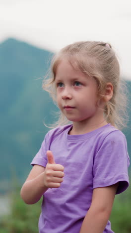 cute little girl with blonde plait shows thumb-up gesture admiring green valley with curving calm river at mountains foot closeup slow motion