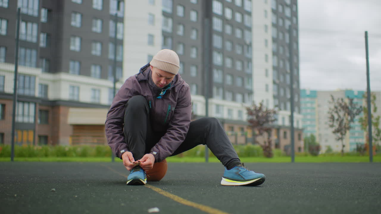 urban court athlete tying sneaker laces beanie clad man sits on basketball adjusting shoes before training amid residential towers under overcast sky focused warmup routine solo preparation for run