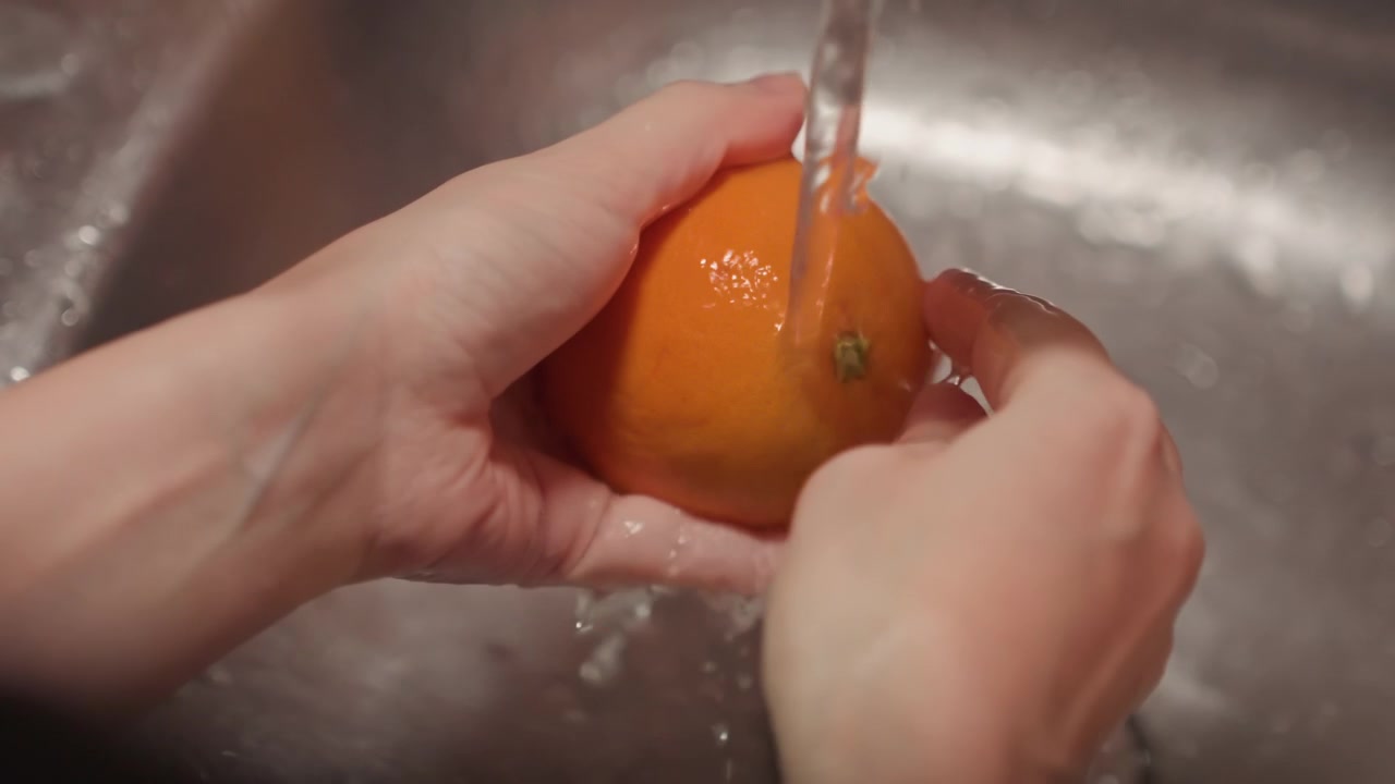 Primer plano de manos lavando una naranja bajo el agua corriente en el fregadero. La fruta se gira y se enjuaga a fondo con la piel visiblemente mojada. El entorno de cocina metálico proporciona contexto