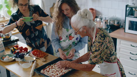 young man taking photos of sister preparing pizza with grandmother in kitchen