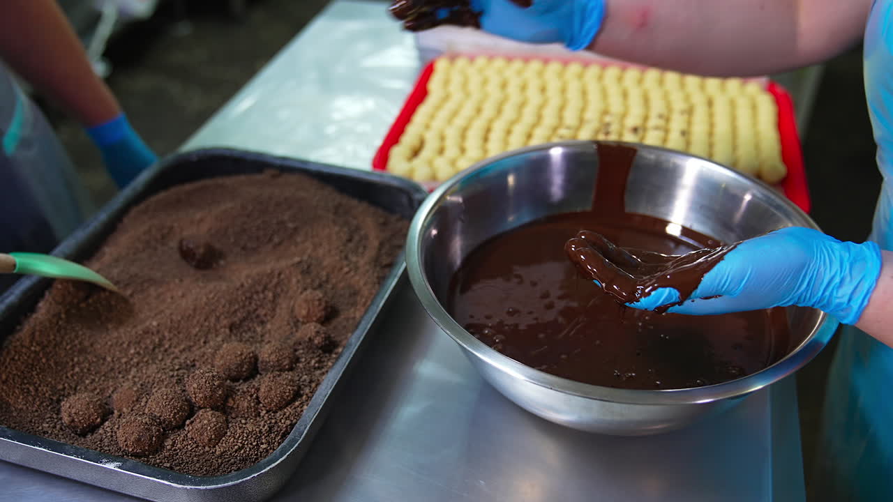 Two confectionery employees making candies by hands. Gloved hands take creamy balls and cover them with chocolate and then with cocoa crumbs.