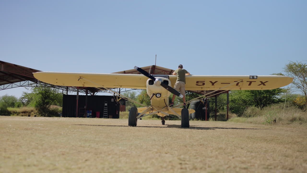 Uomo che sale sull'ala di un piccolo aereo a elica in un campo d'aviazione.