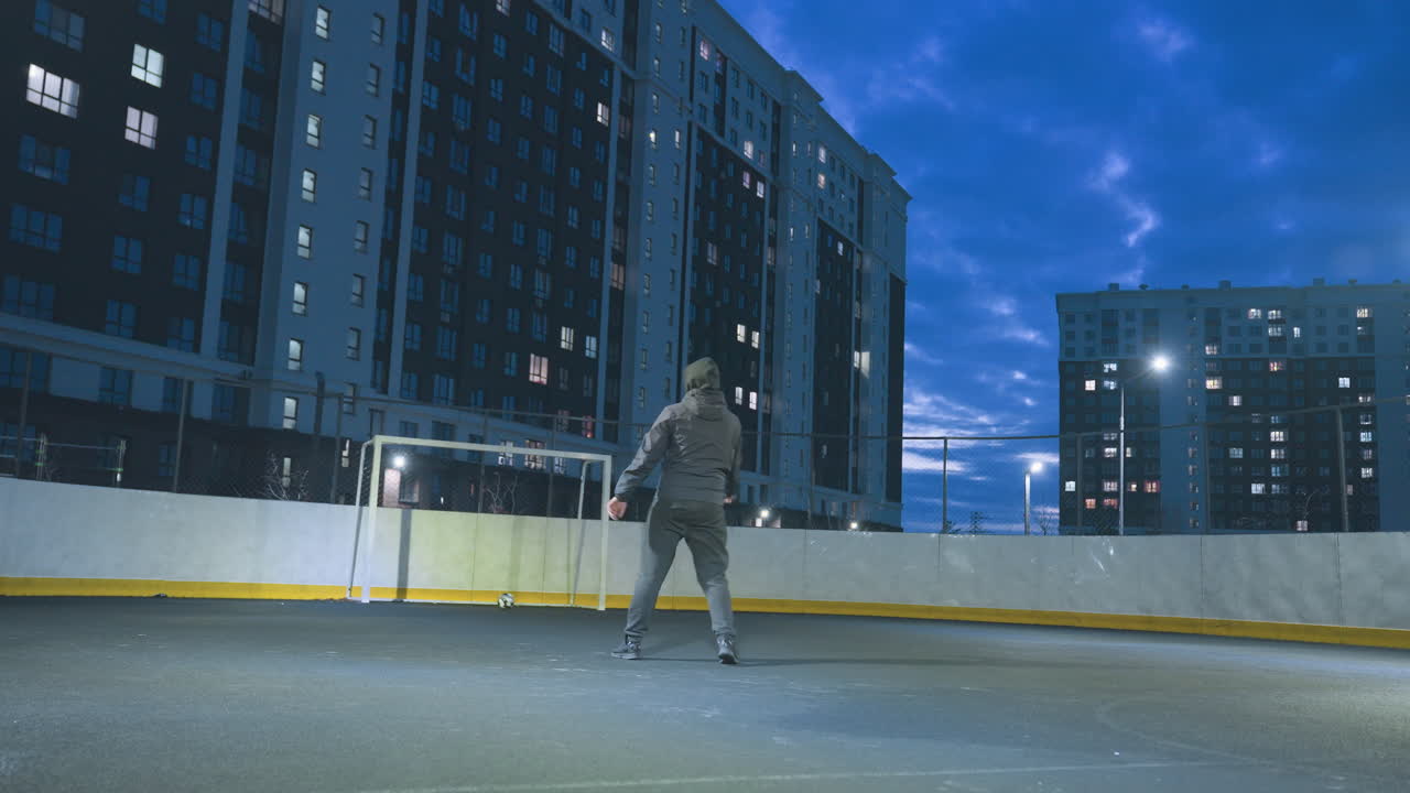 Athlete kicks soccer ball towards goalpost during evening training on urban field under bright floodlights, surrounded by tall residential buildings and deep blue sky