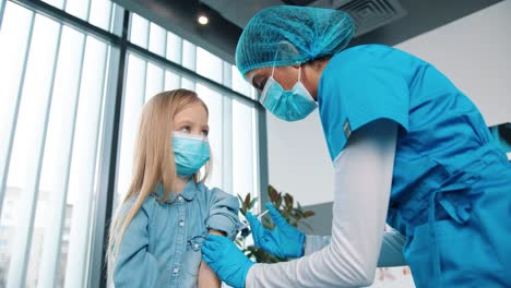 Close-Up-Of-Professional-Healthcare-Specialist-Female-Nurse-In-Protective-Uniform-And-Medical-Mask-Making-Covid-19-Vaccine-Injection-To-Little-Adorable-Kid-Girl-In-Clinic-Lab-Room,-Vaccination-Concept