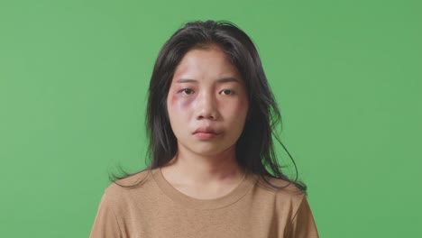 close up of young asian woman victim of violence with bruise on face looking painful into the camera on green screen background in the studio