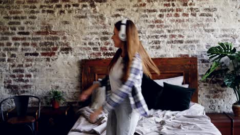 attractive young woman in casual clothes is dancing on bedroom floor and listening to music in headphones. modern loft style apartment and plants are visible.