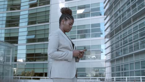 Static-shot-of-a-focused-African-American-businesswoman-standing-among-city-office-buildings-and-using-cell-phone