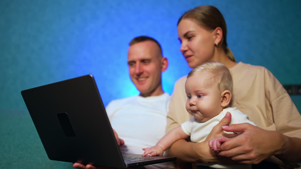 Happy Caucasian parents with lovely blond infant sit on sofa. Smiling family look at laptop in man's hands. Low angle view.
