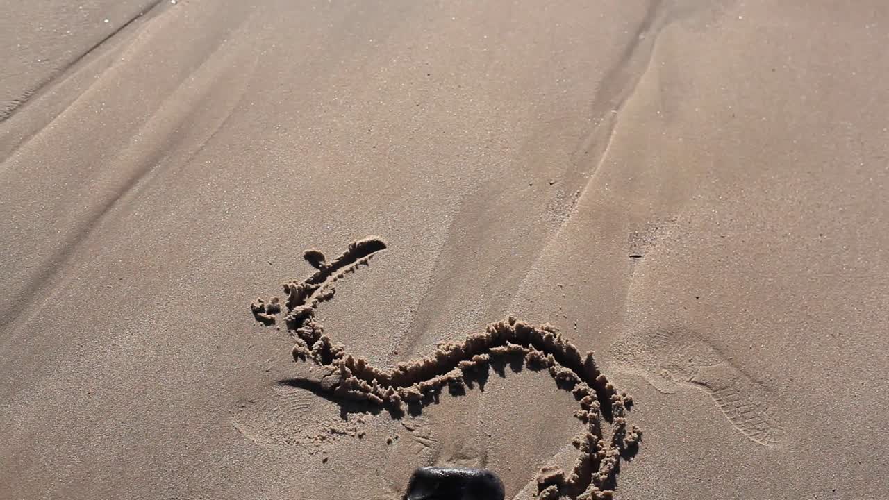 Girl is drawing on a beach in the sand letter S. It is a sunny day. Camera is directed downwards. Girls hair are swaying in front of camera. Filmed in HD