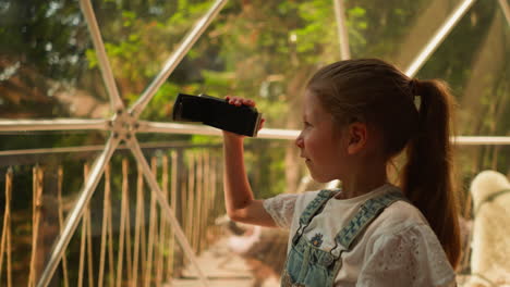 child records shots with camera at glamping. concentrated little girl shoots video through panoramic window on hotel terrace. kid takes shots