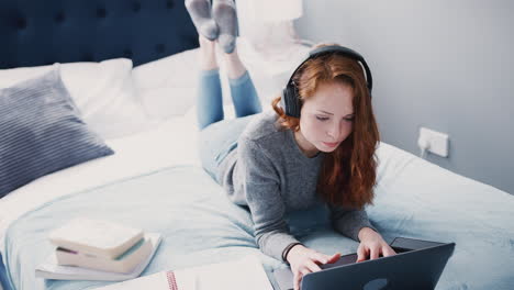 female college student wearing headphones works on bed in shared house with laptop and mobile phone