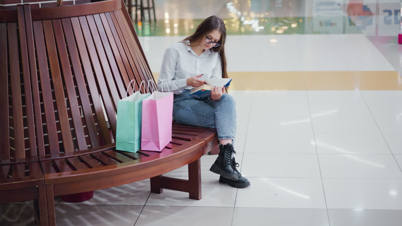 estudiante sentado en un centro comercial bien iluminado con las piernas cruzadas, leyendo un libro con reflejos de luz en los azulejos, bolsas de compras al lado, y un banco de madera moderno que añade a la atmósfera contemporánea